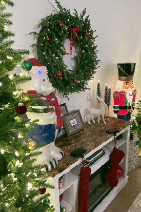 A wreath with lights, ornaments, and a bow hanging on the wall atop a fireplace mantel.